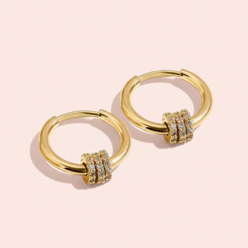 Gold hoop earrings with embedded stones on a light pink background